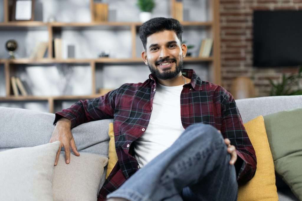 Smiling young middle east male renter or tenant relax on sofa at home, enjoy weekend.